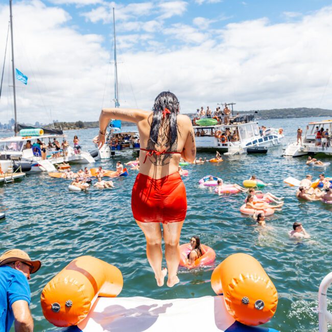 A woman in a red swimsuit jumps off a boat into a body of water adorned with colorful inflatables while numerous people on other boats and in the water around her enjoy the sunny day. The sky is partly cloudy, and the scenic background features more boats and a hilly landscape.