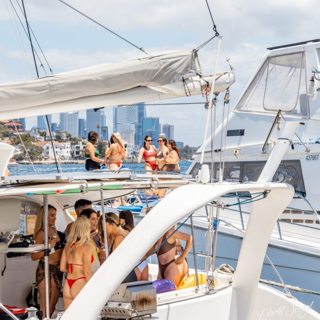 A group of people in swimsuits enjoying a sunny day on a yacht cruise. Some are chatting and taking pictures, while others are relaxing on the deck with refreshing drinks. The background features another boat, distant buildings, and a partly cloudy sky.