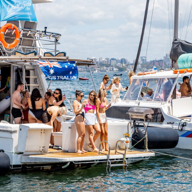 A group of people enjoying a sunny day on a docked boat displaying an Australian flag. Some are dancing, while others relax and chat. Another boat with people is docked nearby, with city skyscrapers and the sparkling water of the harbor visible in the background.