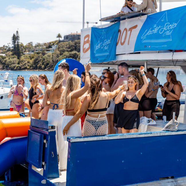 A large group of people in swimwear are gathered on a docked double-decker boat, enjoying a sunny day. Some are dancing, some are socializing with friends, and others are taking photos. The boat has a banner that reads "The Yacht Social Club." Nearby trees and buildings add to the scenic background.