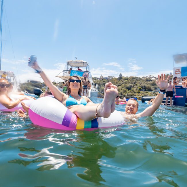 Three people enjoying a sunny day in the water. A woman in the foreground is lounging on a colorful pool float, raising a drink can and stretching out her legs. Behind her, a man and another woman are also relaxing near some boats, savoring the clear, blue waters together.