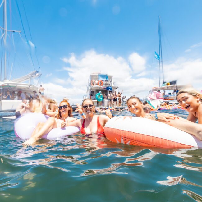 Four people are floating on inflatable pool floats in the ocean, smiling under a sunny sky. Several boats are anchored nearby, and other people are visible enjoying the water and paddleboarding in the background. The scene is lively and festive.