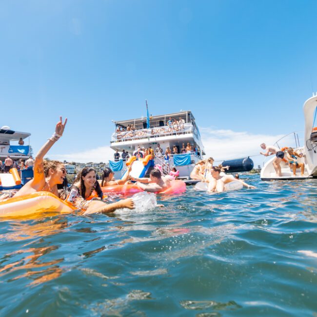 People enjoying a yacht party on the water. Various individuals are floating on inflatable rafts and inner tubes, swimming, and socializing near several luxury yachts. The sky is clear and sunny, creating a festive and lively atmosphere filled with joyful laughter.