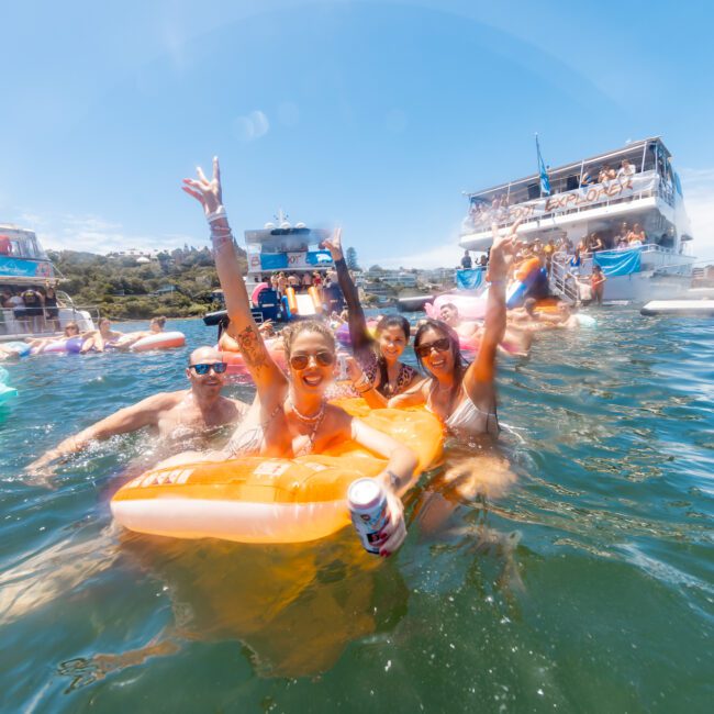 A group of people are enjoying a sunny day in the water, holding floaties and drinks. The background shows multiple boats and other people swimming and relaxing. The atmosphere appears festive and lively, like a floating party. The text "The Yacht Social Club" is visible in the bottom right corner.