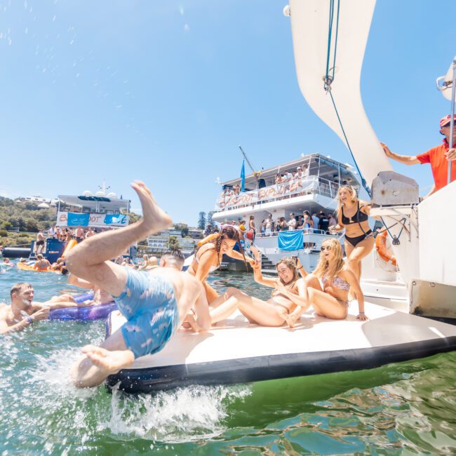 A group of young people relax and sunbathe on a boat's deck while a man in blue swim trunks performs a cannonball into the water. Other boats and people can be seen in the background, enjoying a sunny day on the lake. The scene is lively and cheerful.