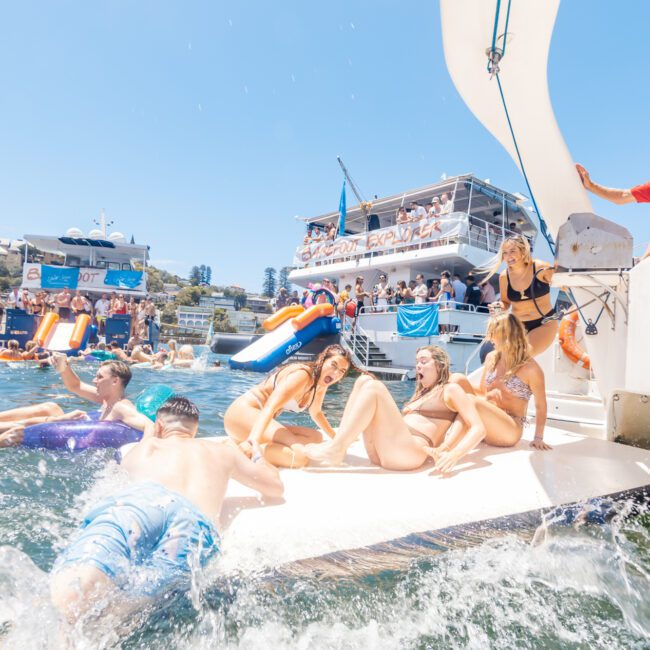 A group of people is enjoying a sunny day on a boat, with some lounging in swimwear on the deck and others playing in the water. A large waterslide extends from the boat, leading to inflatables visible in the background. The sky is clear and blue as everyone relishes under the bright sun.