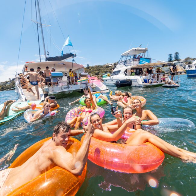 A lively scene of a yacht party on the water with people enjoying the sun and waves in colorful inflatable tubes. Several yachts are in the background, and attendees are swimming, socializing, and having drinks, creating a joyful and festive atmosphere.