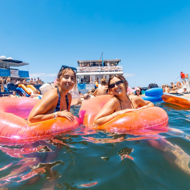 Two people are smiling and lounging on pink inflatable floats in the water, surrounded by numerous other individuals enjoying a sunny day. Several yachts and a floating platform with a slide are in the background, while the clear blue sky enhances the vibrant scene.