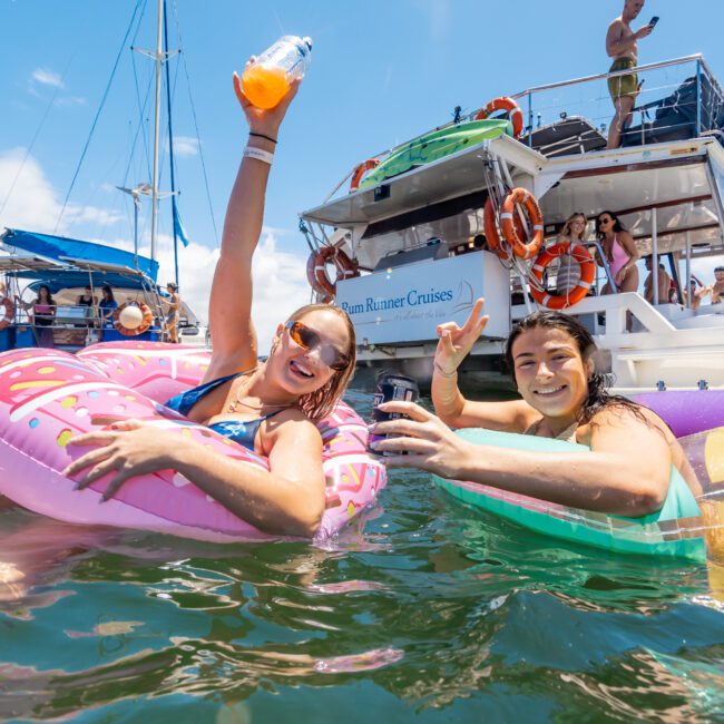 Two people float in the ocean on inflatable tubes, smiling and holding drinks. A boat named "Rum Runner Cruises" is in the background with others on board enjoying the clear, sunny sky. The atmosphere is lively and joyful under the radiant sun.