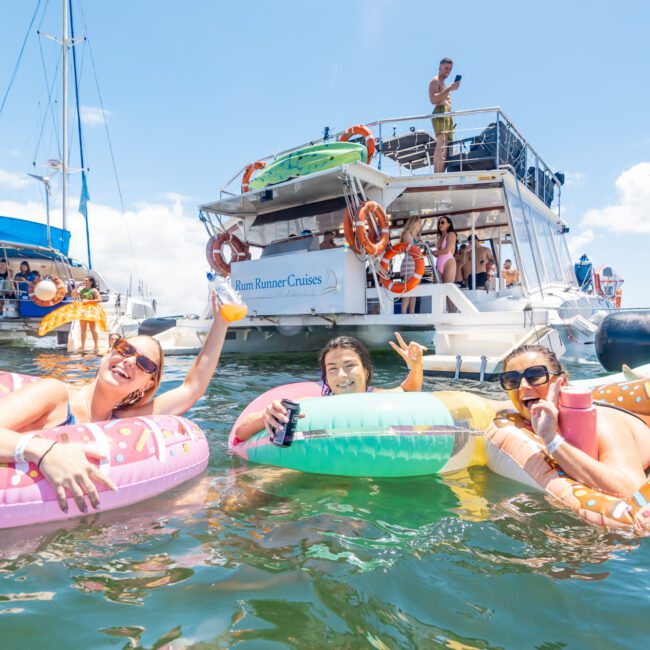 Three people enjoying the water on inflatable rings near a boat named "Rum Runner Cruises." They are smiling and holding drinks. Others are on the boat, some taking pictures, under a clear and sunny sky with another sailboat visible in the background.