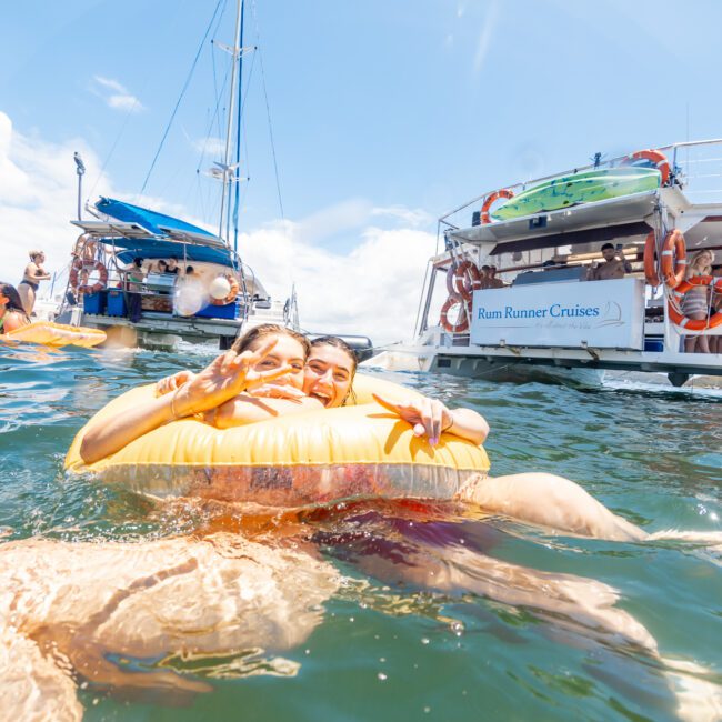 Two people float on an orange inflatable ring in clear blue water, smiling and making peace signs. Behind them, colorful boats with people aboard and a banner reading "Rum Runner Cruises" are anchored. The sky is bright and sunny, enhancing the festive atmosphere.