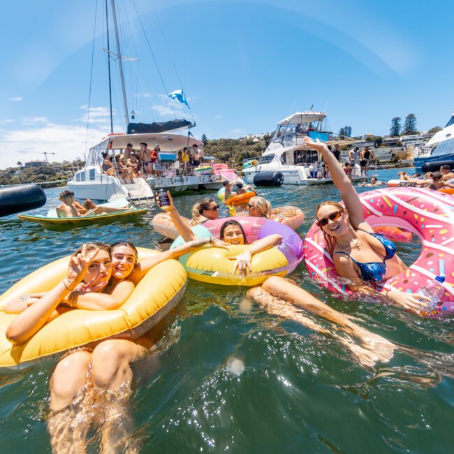 A group of people are enjoying a sunny day on a lake, floating on inflatable pool tubes, including a donut-shaped one. In the background, several boats are anchored. People are swimming, laughing, and having fun in the water under the "Yacht Social Club" banner.