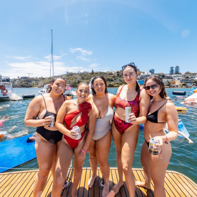 A group of five smiling women in swimsuits stand on the deck of a boat, holding drinks. Behind them, people are swimming and floating on inflatables in the water. Multiple boats and a rocky shoreline are visible in the background. The scene at The Yacht Social Club Sydney Boat Hire is sunny and cheerful.