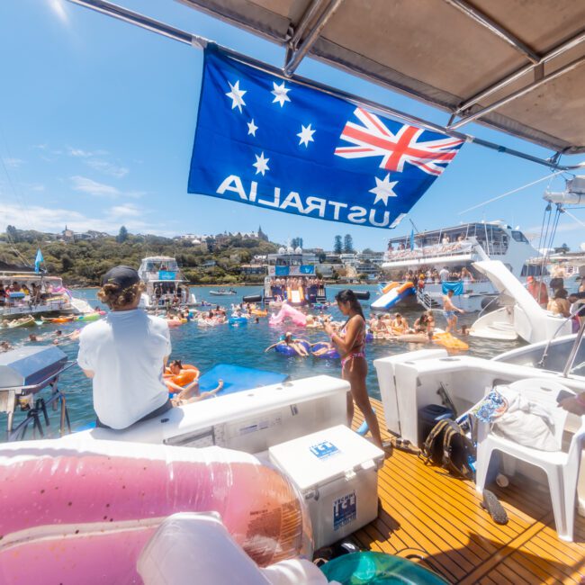 A lively scene of people enjoying a boat party, with several boats anchored close together in a sunny bay. An Australian flag is visible, and people are swimming in the water, lounging on inflatable floats, flying kites, and socializing on the boats.