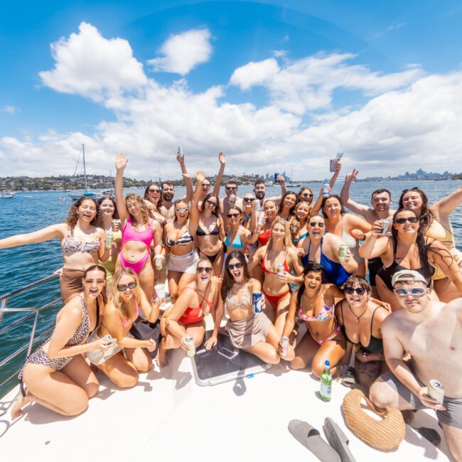 A large group of people in vibrant swimwear are gathered on a boat, enjoying a sunny day on the water. They are smiling, holding drinks, and some have their arms raised in celebration. The blue sky with white clouds and distant sailboats enhance the festive atmosphere in the background.