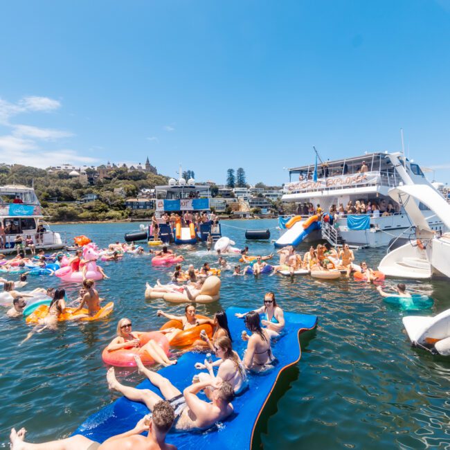 People are enjoying a sunny day on the water, lounging on a large blue floating mat and inflatable floaties. Several boats from The Yacht Social Club Sydney Boat Hire are anchored nearby, with others standing or sitting on deck, all having a good time. The background shows a shoreline with greenery and buildings.