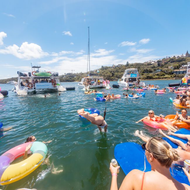 People are enjoying a sunny day on the water in an inlet. Some are swimming while others float on colorful rafts. Boats are anchored nearby, and the background features a hillside with trees and even a charming building. The atmosphere is lively and festive, perfect for an enjoyable summer day.