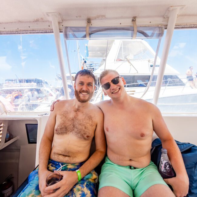 Two smiling men in swim trunks sit closely together on a boat with their arms around each other. The man on the left has a drink in hand, while the man on the right sports sunglasses. In the background, other boats and people are enjoying a sunny day of festivities on the water.