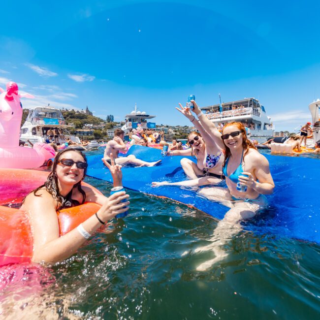 People are enjoying a sunny day at a yacht party, floating in the water with colorful inflatables, including a pink unicorn and a large pink ring. Some attendees are holding drinks and smiling at the camera while upbeat music plays, creating an unforgettable ambiance with boats anchored in the background.