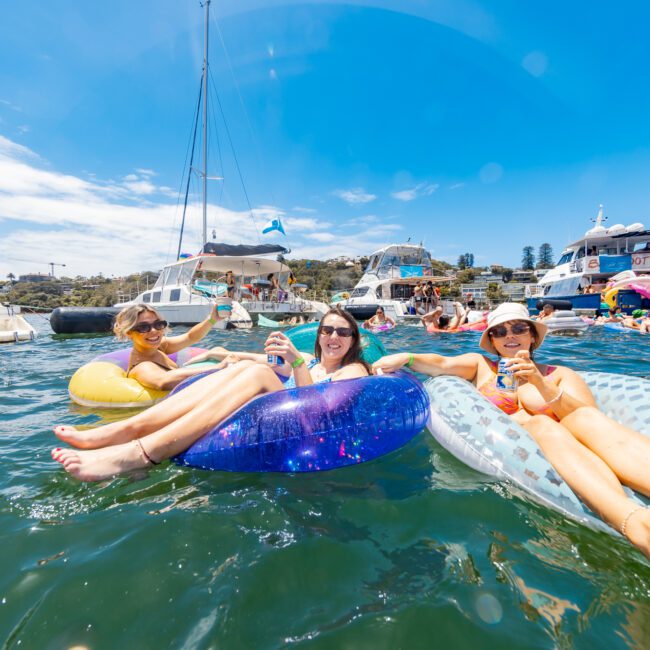 Three people float on inflatable tubes in a sunny, clear, and lively water setting, each holding a can. Boats and more people are visible in the background, suggesting a joyful recreational gathering on the water. Text reads "Yacht Social Club.