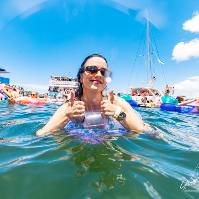 A woman in sunglasses smiles and gives two thumbs up while in the water. She is surrounded by others on inflatables, and boats are visible in the background under a clear blue sky. The "Global Social" logo is at the bottom right corner of the image, capturing a perfect summer vibe.