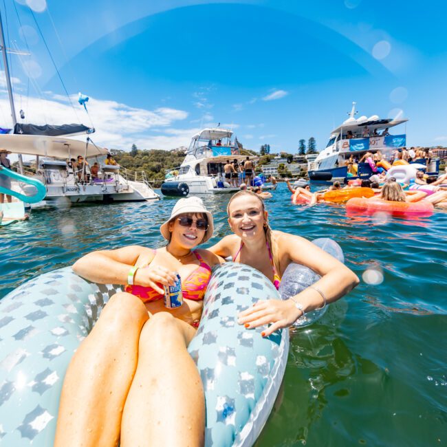 Two people in swimsuits relax on inflatable pool floats in a sunny, crowded waterway with boats and other floaters in the background. One holds a beverage can and smiles at the camera. The clear sky adds to the festive and lively atmosphere, making it an idyllic summer day.