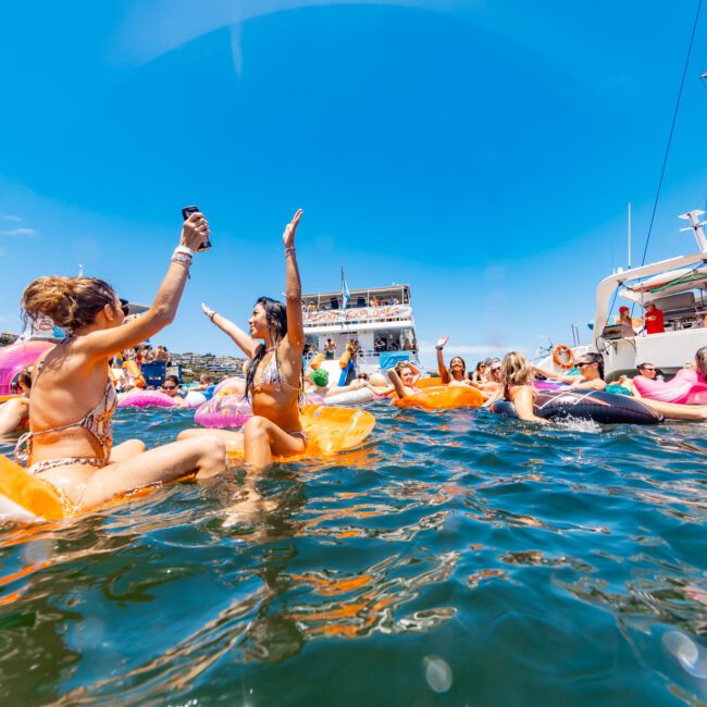 People are floating on inflatable tubes and rafts in the water, having fun and raising their hands. A boat and a few yachts are in the background under a clear blue sky. The scene is sunny and festive, suggesting a beach party atmosphere.