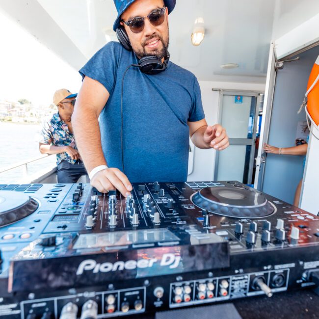 A DJ stands on an outdoor deck under a sunny sky, wearing sunglasses, a blue hat, and headphones around his neck while operating a Pioneer DJ controller. In the background, another person in a patterned shirt and hat adds to the vibrant atmosphere.