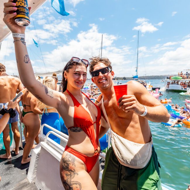 A woman in a red swimsuit and sunglasses, with a tattoo on her left thigh, and a man in green shorts with a fanny pack, holding drinks and posing on a boat. In the background, people are partying on nearby yachts under the bright sun.