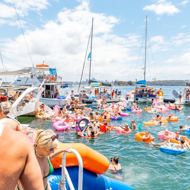 A lively scene of people enjoying a day on the water. Individuals float on colorful inflatable tubes and rafts near several anchored boats. Various people seen in swimwear, with a clear sky and flags waving in the breeze. A serene waterfront enhances the inviting ambiance.