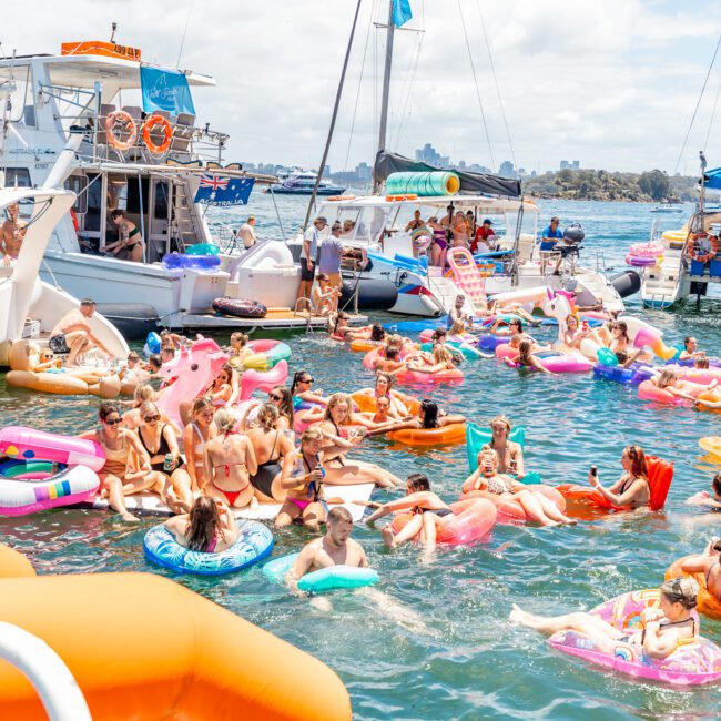 A lively scene of people enjoying themselves at a boat party on a sunny day. Many are floating on vibrant inflatables in the water, while some remain on nearby boats. The backdrop features a clear sky, calm waters, and a distant shoreline with lush vegetation.