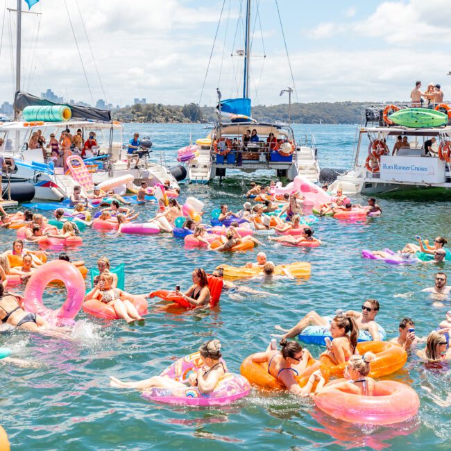 A large group of people are enjoying a sunny day on the water, floating on colorful inflatable rings and loungers. Several boats from The Yacht Social Club are anchored nearby, with more people on board. The scene is lively and festive, set against a backdrop of distant shores.