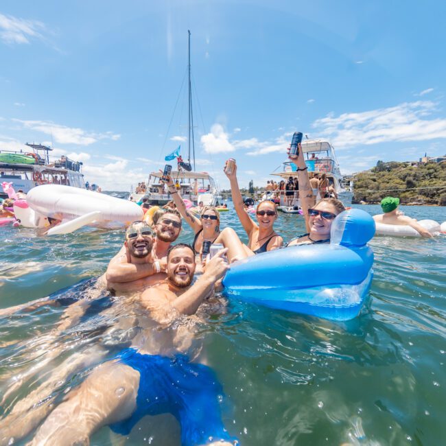 A group of people enjoying a sunny day in the water, surrounded by colorful inflatable floats with boats in the background. They are smiling and raising their drinks, creating a lively and festive atmosphere. The sky is clear, and tall trees are visible in the distance.