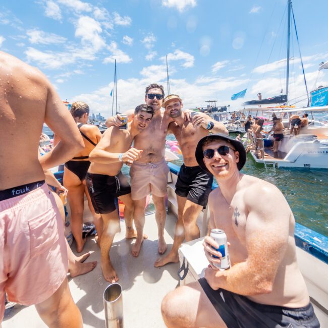 Four men in swimwear pose cheerfully on a crowded boat in a sunny, open water setting filled with boats and people. One man, holding a can, kneels in front while the other three stand closely together, smiling. It’s a lively, festive atmosphere reminiscent of summer beach parties.