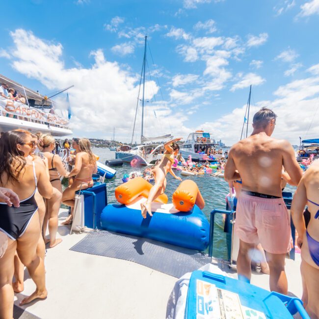 A lively yacht party with numerous people in swimsuits congregating on boats and inflatables. The scene includes a woman in an orange bikini walking up an inflatable slide, with others mingling, enjoying drinks, and the sunny weather. Branded "The Yacht Social Club," it epitomizes a perfect summer escape.