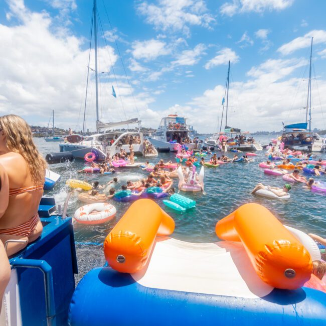 A lively scene of a yacht social gathering in sunny weather. People in swimsuits are floating on various inflatables in the water, surrounded by several anchored boats. An inflatable slide leads into the water from a yacht hosting The Yacht Social Club Event Boat Charters.