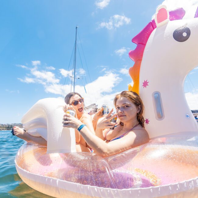 Two people are enjoying a sunny day on the water, relaxing on an inflatable unicorn float. They are holding cans and smiling at the camera. A sailboat is visible in the background against a clear blue sky. The scene is cheerful, with laughter echoing across the vibrant landscape.