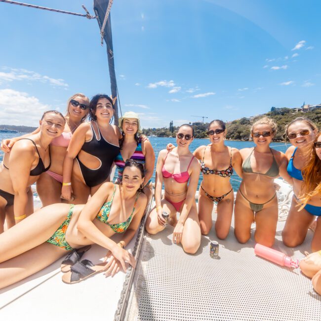 A group of women in vibrant swimwear pose together on a boat under a sunny blue sky. They are smiling and enjoying the day, with a scenic view of the water and shoreline enhancing the background.