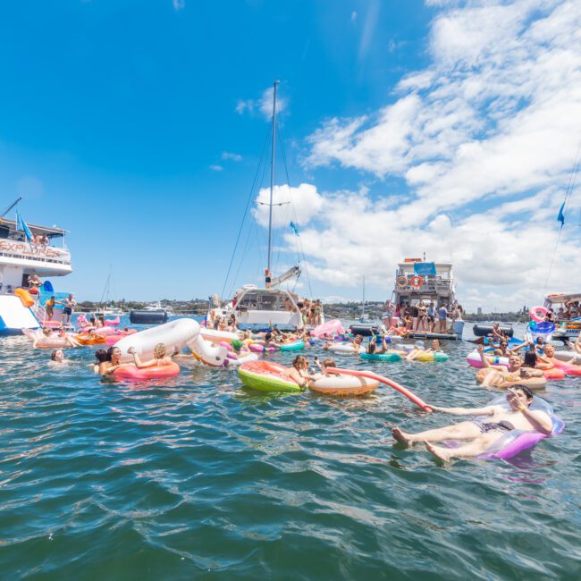 A lively scene on the water with people enjoying a sunny day on inflatable floats of various shapes and colors. Nearby, several boats are anchored, including a vibrant party yacht. The sky is bright blue with scattered clouds, creating a festive atmosphere.