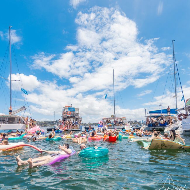 People are relaxing and floating in colorful inflatables on a sunny day in the water near several boats. The sky is blue with some white clouds, and the scene is vibrant and festive with a sense of tranquility. The calm water reflects the bright sky above.