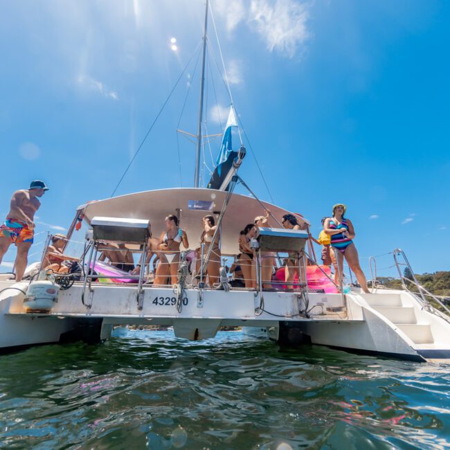 A group of people enjoying a sunny day on a catamaran. Some are on the deck, while others are preparing to get into the water for snorkeling. The sky is clear and blue, and other boats are visible nearby. The scene is lively and relaxed.