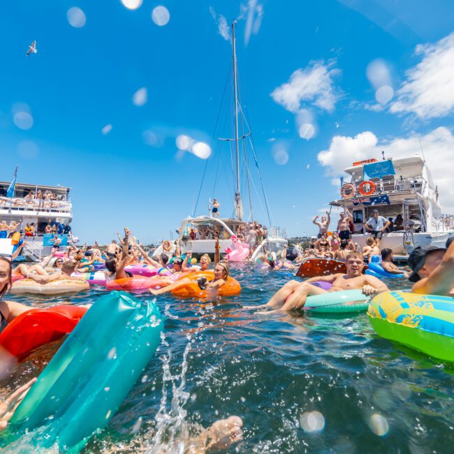 A lively party scene on the water features numerous people enjoying themselves on colorful floaties. Boats are anchored nearby, and attendees are dancing and mingling in swimsuits. A clear blue sky with a small airplane banner in the background adds to the vibrant atmosphere.