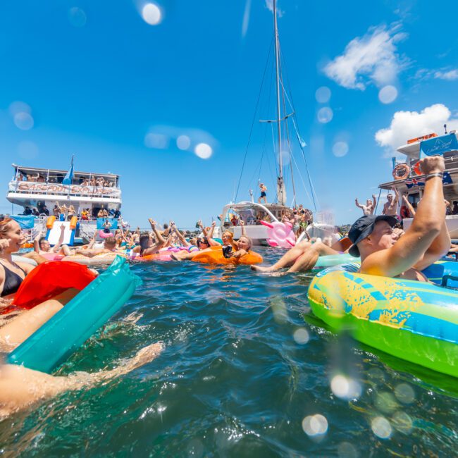 People enjoying a vibrant party on the water with numerous colorful floaties and boats in the background. The scene depicts a sunny day with clear blue skies and multiple people raising their hands, having fun in crystal-clear waters.