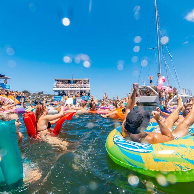 A lively crowd enjoys a sunny day on a lake, floating on inflatable tubes and rafts near docked boats. The clear blue sky and splashes of water create a vibrant and festive atmosphere. A yacht, along with paddle boarders navigating nearby, adds to the scenic backdrop.