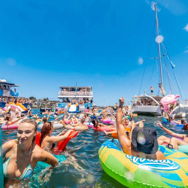 A large group of people enjoying a sunny day in the water, floating on colorful inflatables near several anchored boats. The sky is clear and blue, and the atmosphere is lively and festive with people smiling and having fun, creating unforgettable summer memories. A branded logo is visible in the bottom corner.