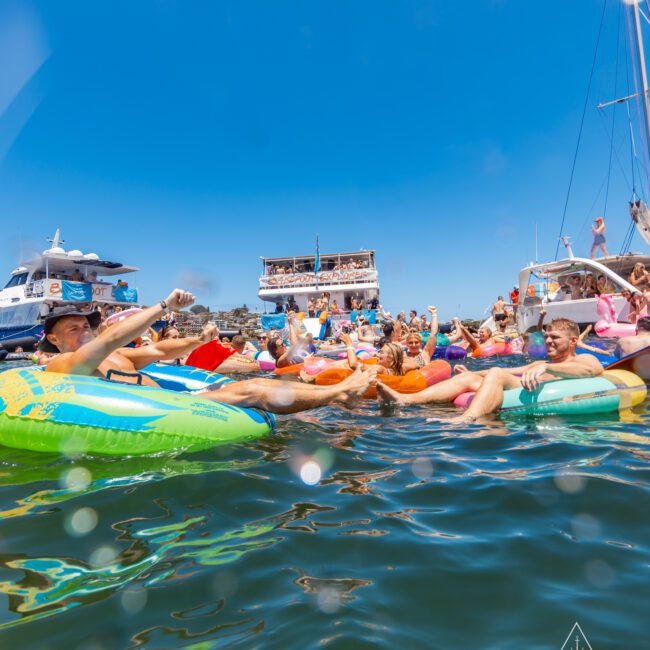 A vibrant scene of people lounging on colorful inflatable floats in the water, with boats and a party barge in the background. The clear blue sky is dotted with a seagull in flight. The atmosphere is festive and relaxed, ideal for an unforgettable social gathering on the water.