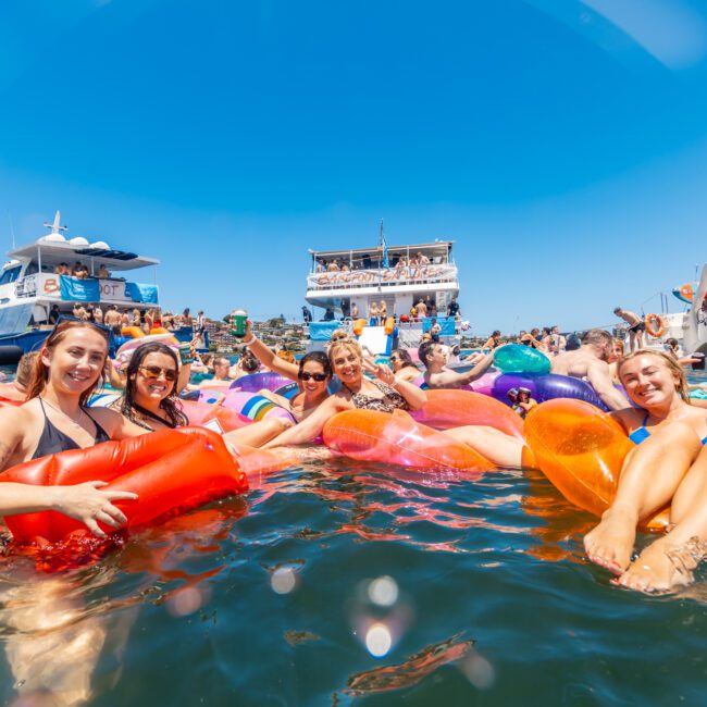 A large group of people enjoys a sunny day floating on colorful inflatables in the water, with multiple boats anchored behind them. Everyone appears to be having a blast as they smile and relax under the vibrant sun. The scene is lively and colorful beneath a clear blue sky.