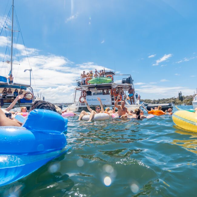 A lively scene on the water, with people in colorful inflatable rafts enjoying a sunny day. Two boats are anchored near each other, with more people on board waving and socializing under the clear sky and bright sunshine. The logo "Yacht Social Club" is proudly visible.