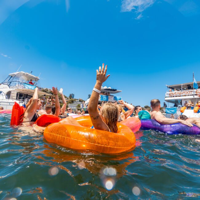 People are enjoying a sunny day at the lake, floating on colorful inflatable rings and rafts, while kayakers paddle nearby. Boats are seen in the background, with one displaying a banner for "Castaway Paddles." Everyone appears to be relaxed, with some waving hands and others lounging.