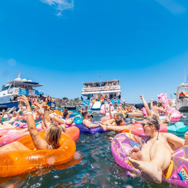 People are enjoying a vibrant and festive time in the water, floating on colorful inflatable tubes and pool toys, surrounded by boats on a sunny day. The lively atmosphere is accentuated by clear blue skies and cheerful inflatables, adding to the summery celebration.